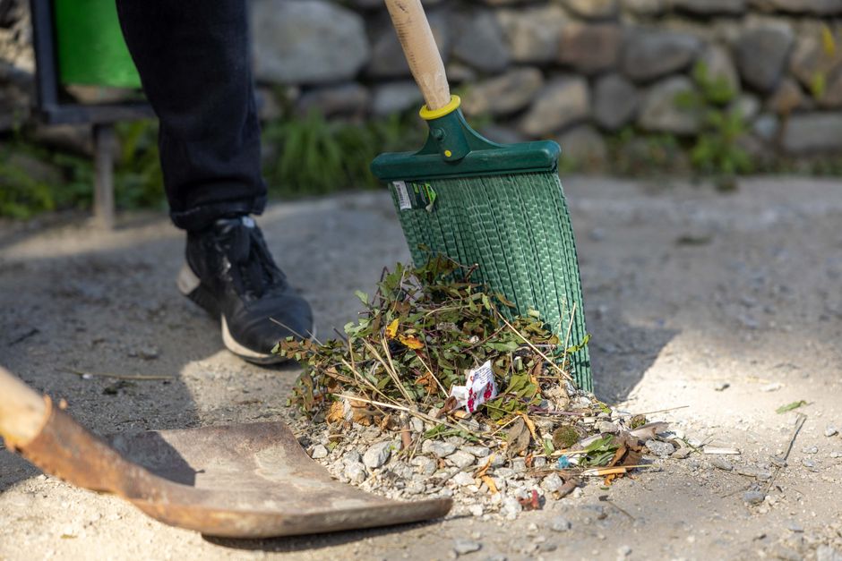 A person wearing black shoes and dark clothing is using a green garden rake to clear leaves, twigs, and small branches from a gravel pathway in a garden. The rake is inserted into a pile of organic wa