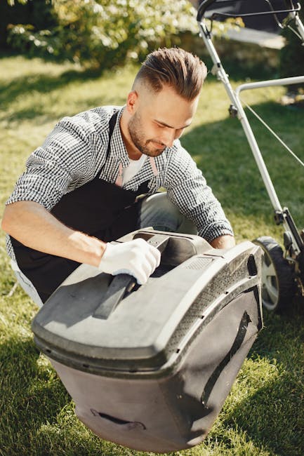 A man with short, styled hair and a trimmed beard is gardening outdoors on a sunny day, wearing a checkered shirt and black apron. He is kneeling on lush green grass and inspecting a large, black gard
