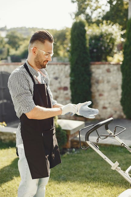 A man with short, styled hair and a trimmed beard is gardening outdoors on a sunny day, wearing a checkered shirt and black apron. He is kneeling on lush green grass and inspecting a large, black gard