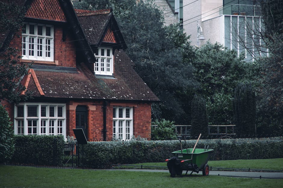 A traditional red-brick house with a sloped, tiled roof and white-framed windows is situated within a landscaped garden. The garden features a neatly trimmed hedge bordering a paved pathway, with a lu